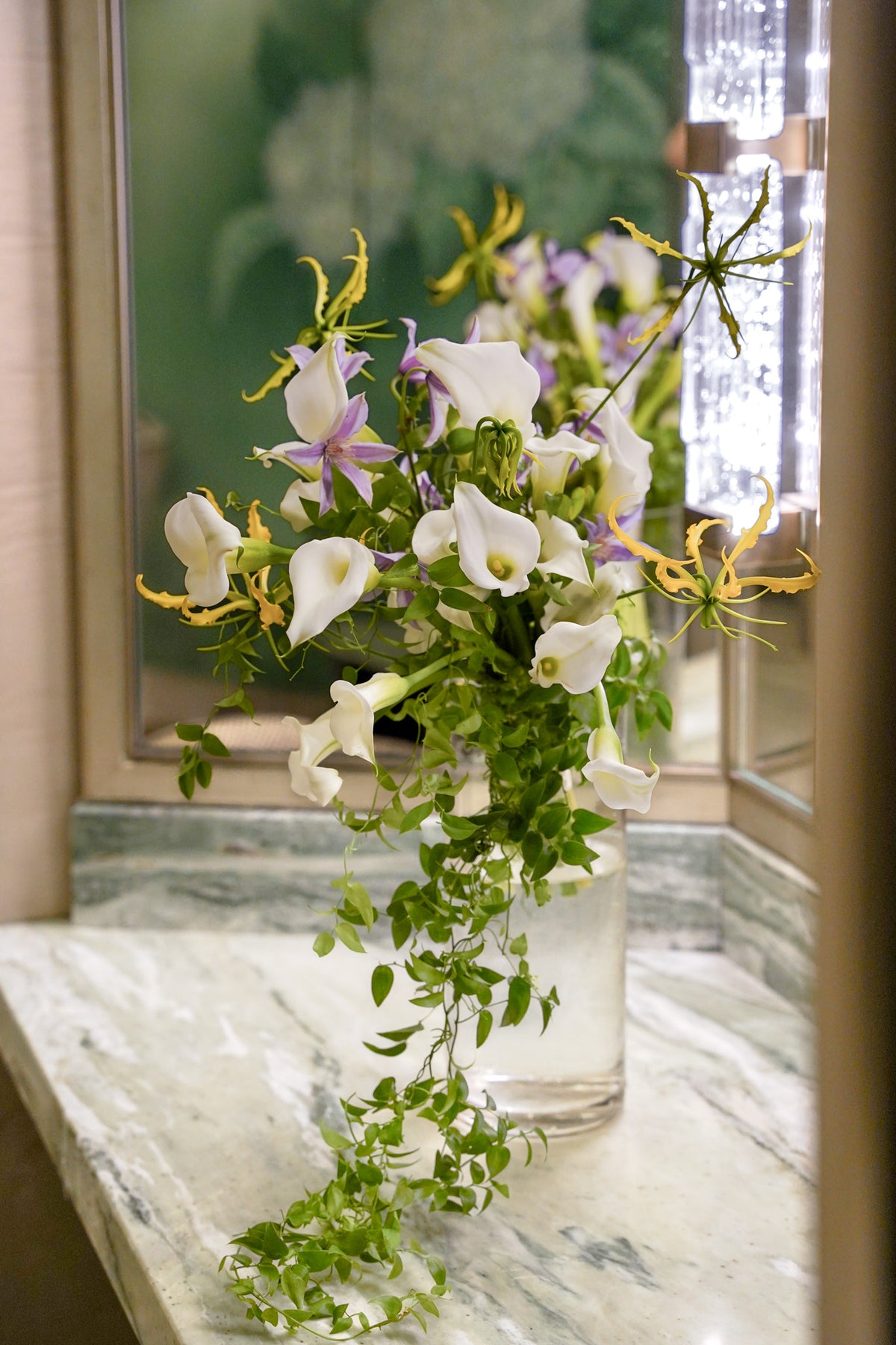 A floral arrangement of white calla lilies, purple irises, and yellow gladiolus in a clear glass vase, displayed on a marble windowsill.